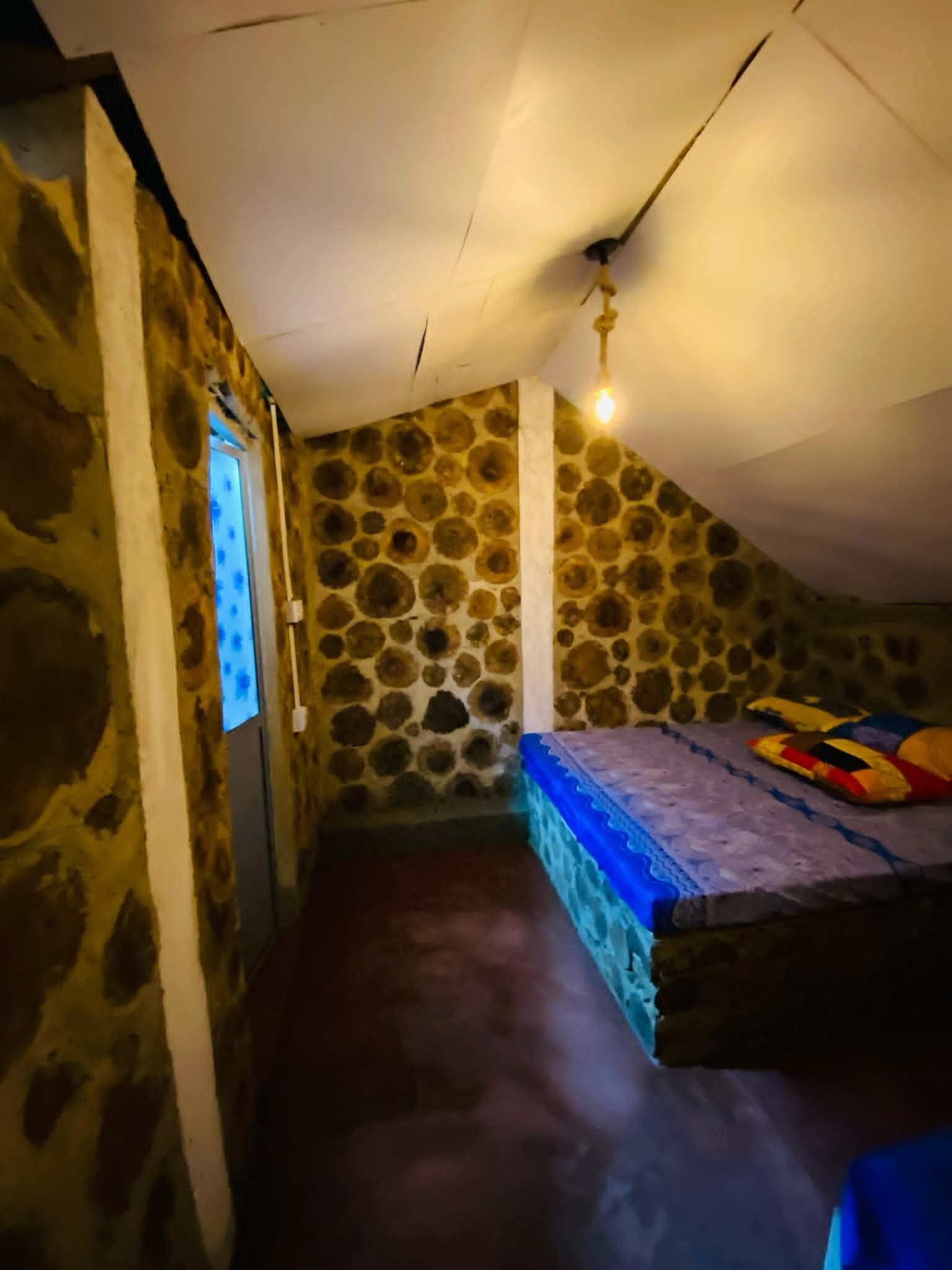 A small, dimly lit room with a sloped ceiling, a single bed with a colorful blanket, and a stone-patterned wall.