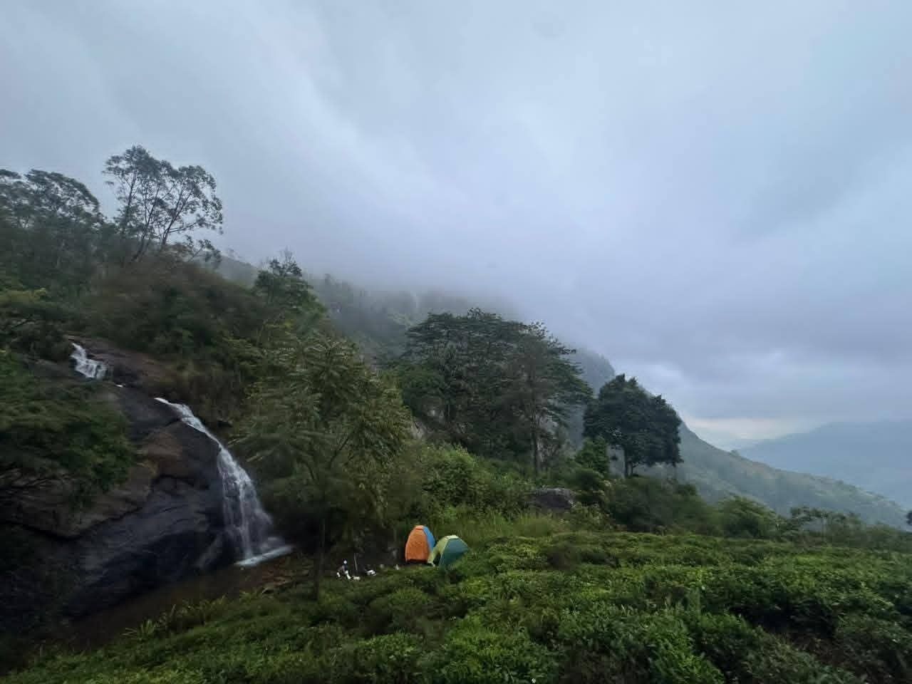 A waterfall cascading down a rocky slope surrounded by lush greenery and a tent set up nearby.
