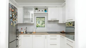 Eine moderne Küche mit weißen Schränken, einem Edelstahl-Kühlschrank, einem Fenster mit Blick ins Grüne und einem eingebauten Backofen.
