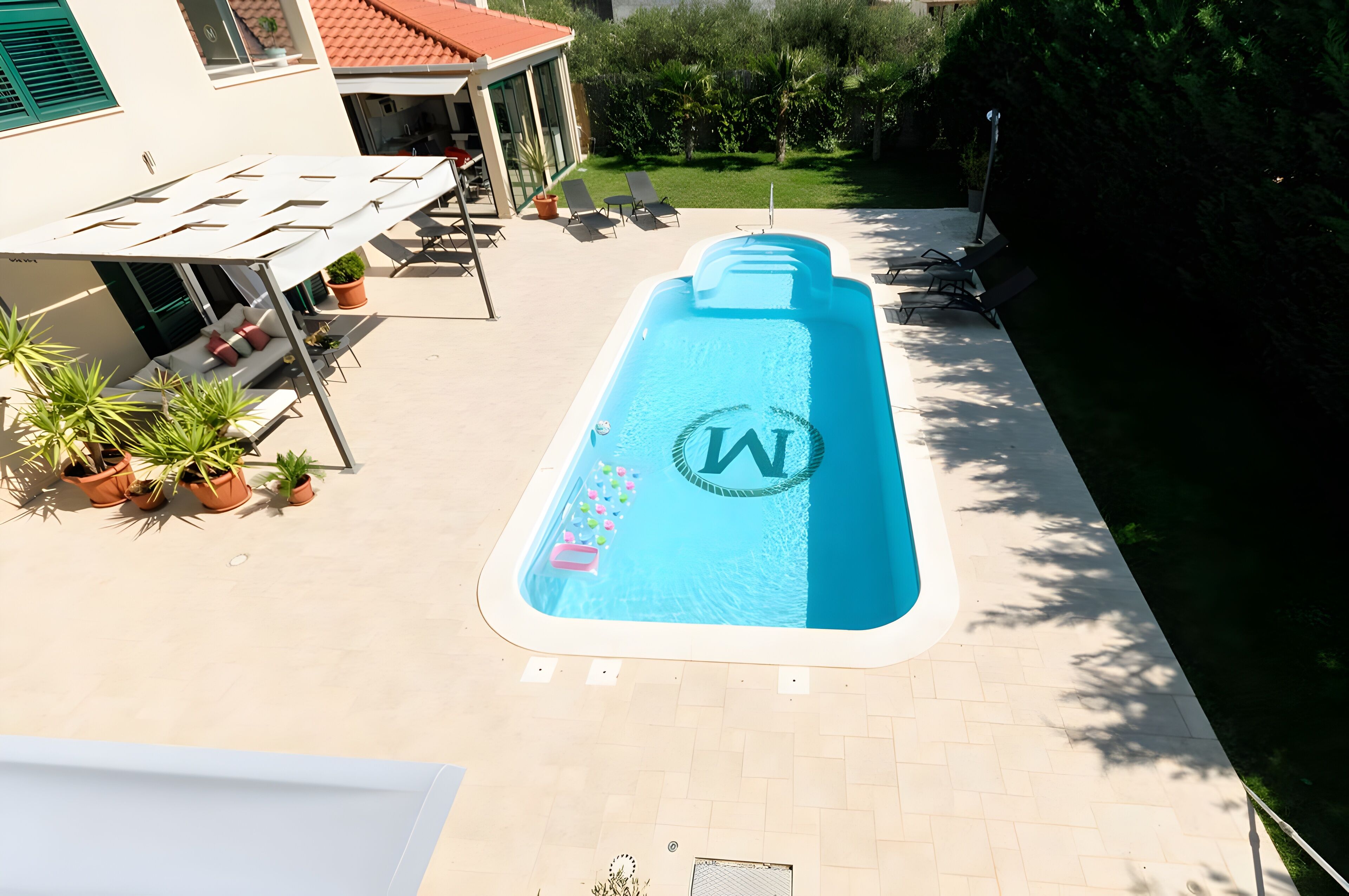 Una piscina con un logotipo, rodeada de plantas en macetas, asientos exteriores y un edificio con techo rojo.