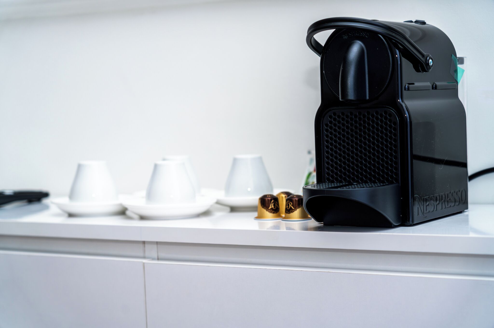 A kitchen counter with a coffee machine, three white cups, and two gold capsules.