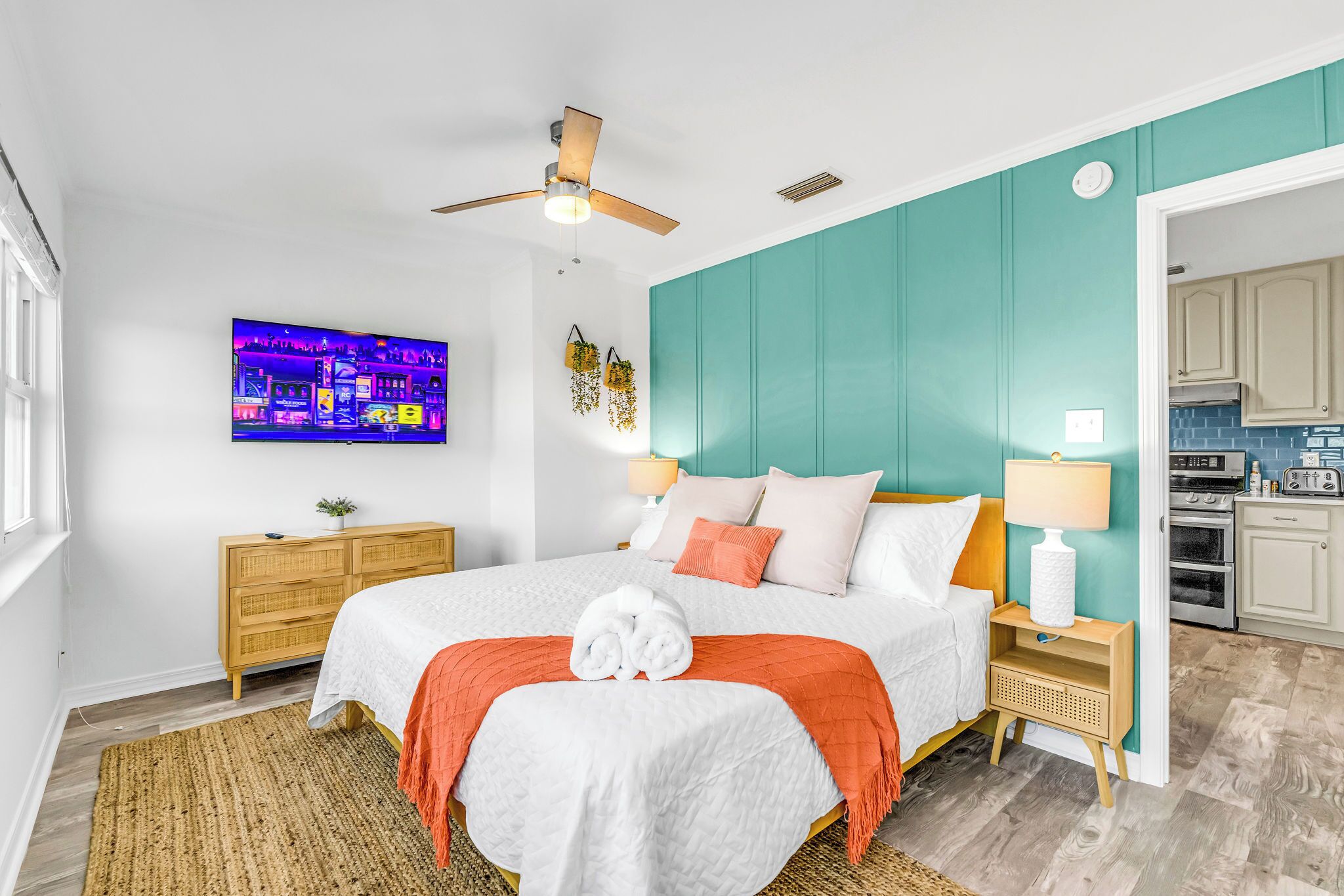 A modern bedroom with a bed, a ceiling fan, a TV, a wooden dresser, and a kitchen area visible through an open door.