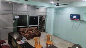 A room with two ceiling fans, a sofa, two chairs, a TV mounted on the wall, and a table with items on it. - Shree Radhe Krishna Guest House (Jhansi)