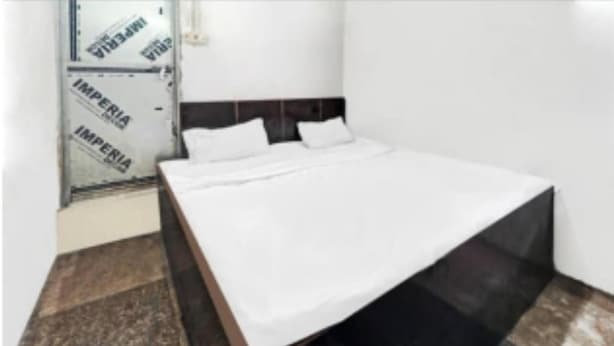 A bed with a white mattress and two pillows, a dark headboard, and a tiled floor with a mix of brown and beige tiles. - Shree Radhe Krishna Guest House (Jhansi)