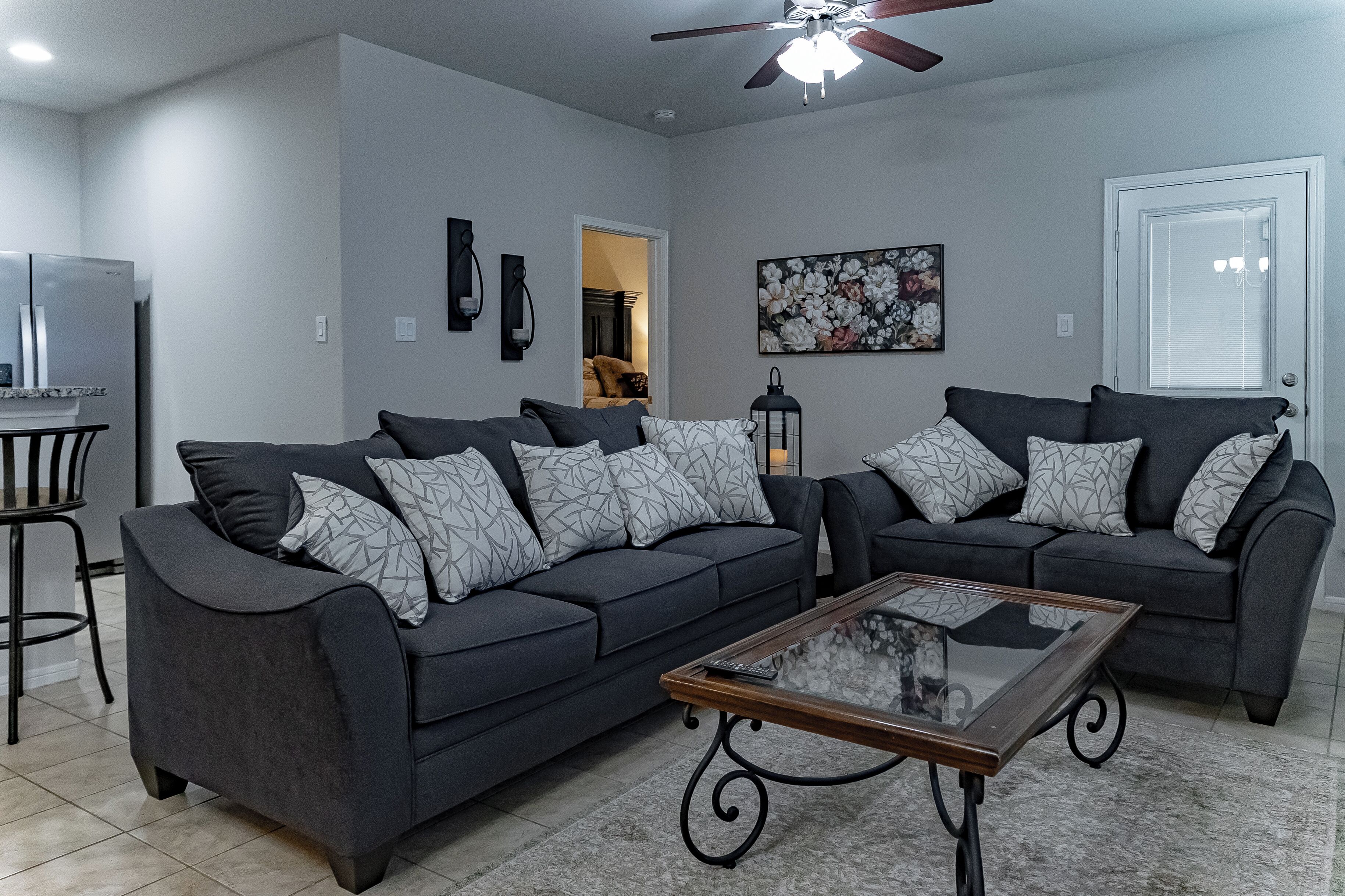 A modern living room with two dark grey sofas, a glass coffee table, and a ceiling fan.