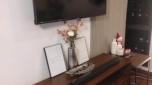 Een moderne woonkamer met een wandgemonteerde televisie, een houten entertainmentmeubel, een decoratieve vaas, ingelijste inspirerende citaten en een uitzicht op de stad 's nachts.