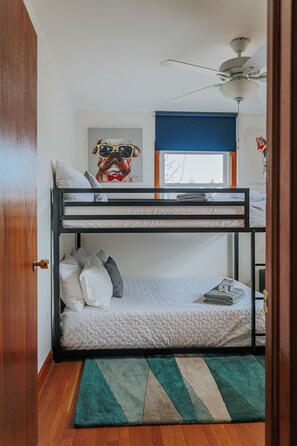 A bunk bed room with a ceiling fan, a framed picture on the wall, and a window with a view of trees.