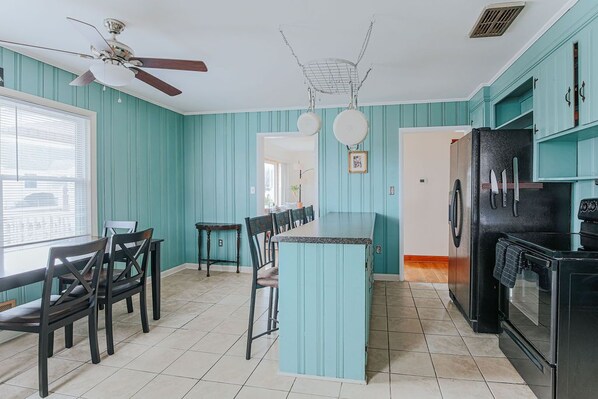 A kitchen with teal walls, a central island, a ceiling fan, a refrigerator, and a dining area with chairs and a table.