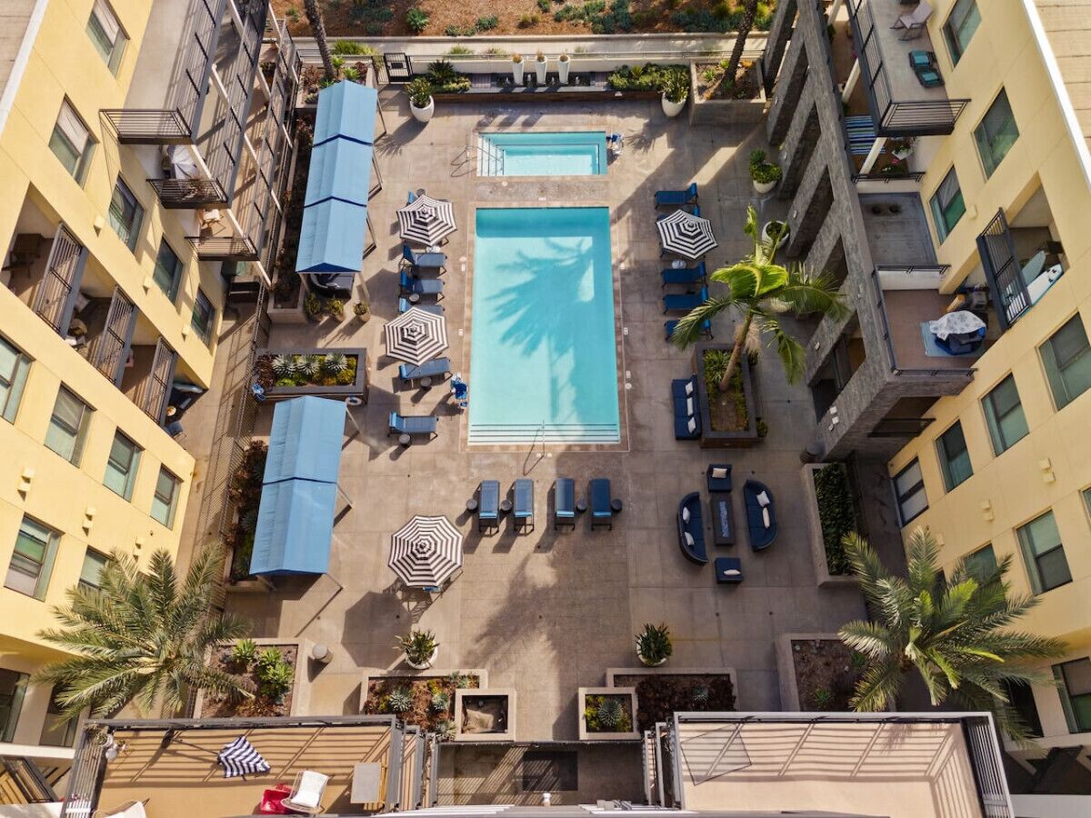An aerial view of a hotel with a central swimming pool surrounded by lounge chairs and umbrellas, with balconies on either side.