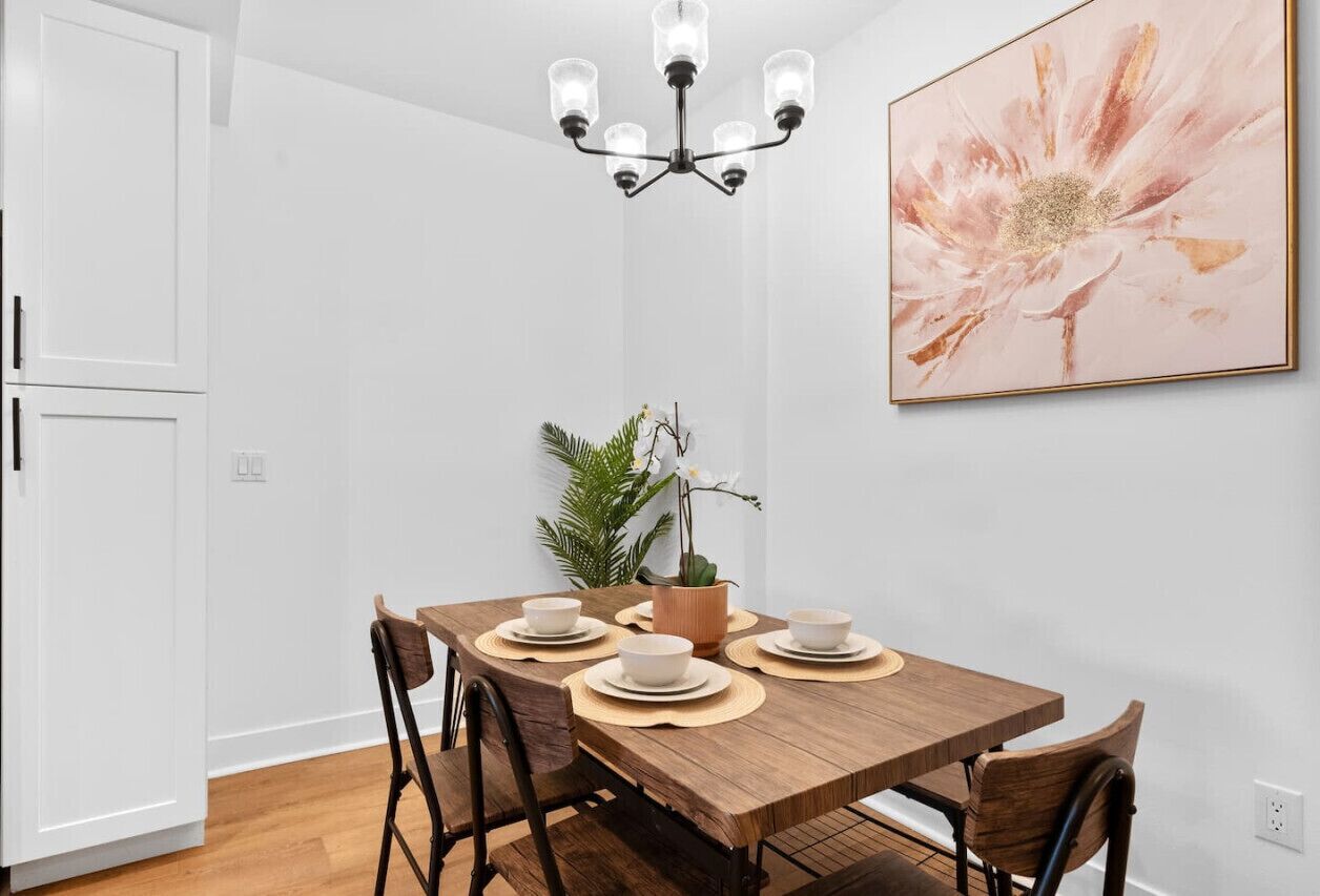 A dining area with a wooden table, four chairs, a chandelier, a potted plant, and a framed floral artwork on the wall.