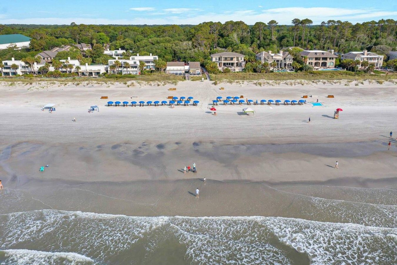 A beach with umbrellas and people, a sandy shore, and houses in the background.