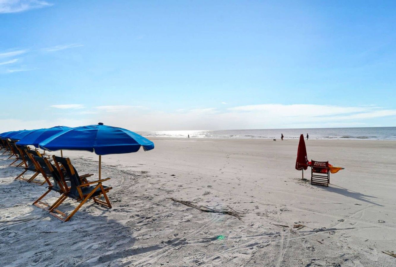 A beach with rows of blue umbrellas and wooden chairs, a lifeguard stand, and a clear view of the ocean.