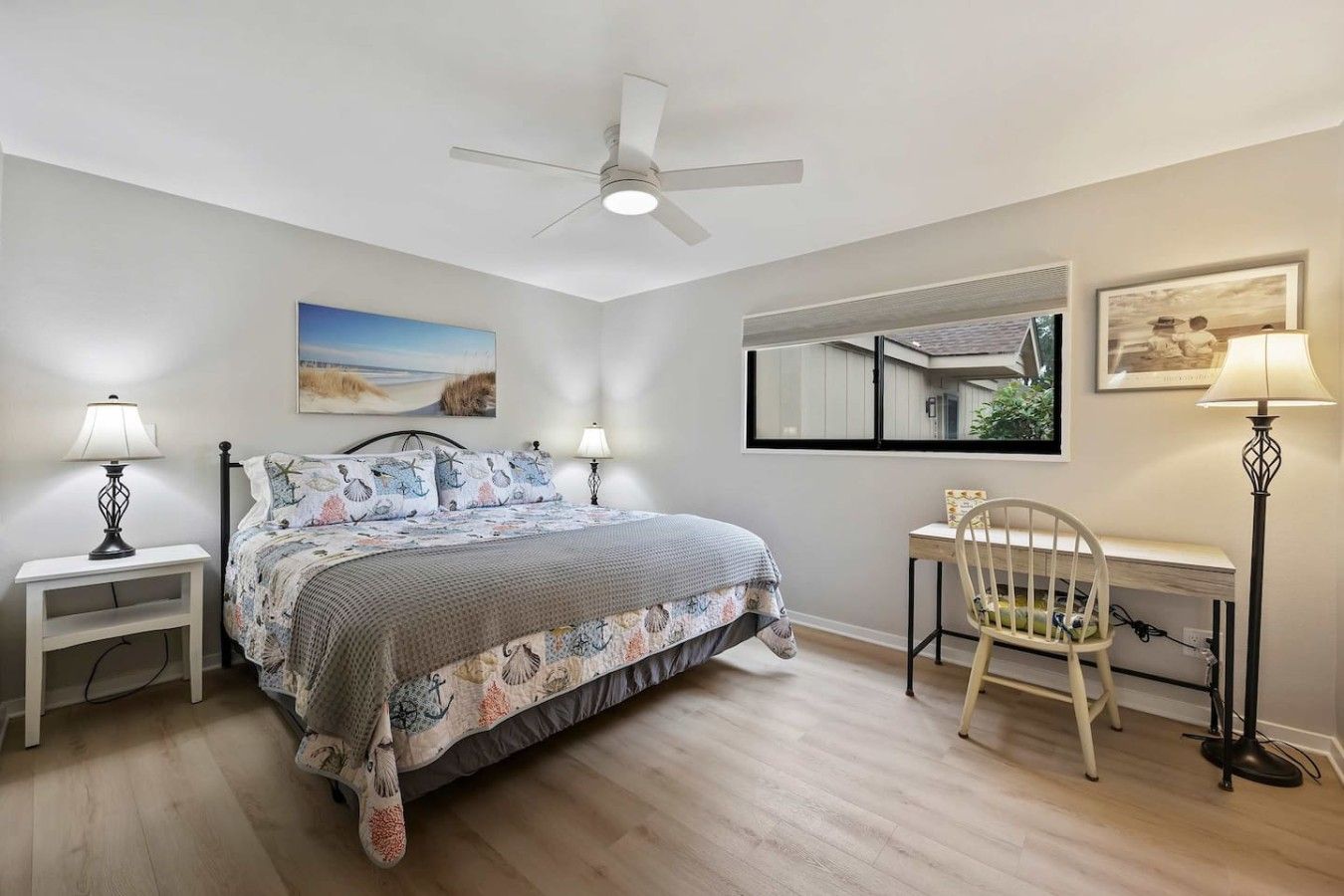 A bedroom with a bed, bedside tables, a desk, a chair, a ceiling fan, and artwork on the walls.