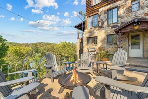 A patio with Adirondack chairs, a fire pit, and a stone building with a view of the forest.