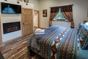 Een slaapkamer met een open haard, een bed met beddengoed met een patroon, een televisie en een raam met gordijnen.