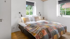 Une chambre à coucher avec un lit, deux fenêtres, un plafond en bois et une vue sur la verdure à l’extérieur.