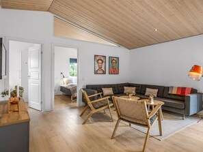 A modern living room with a wooden ceiling, a grey sofa, two armchairs, a coffee table, and a dining area with a wooden table and chairs.