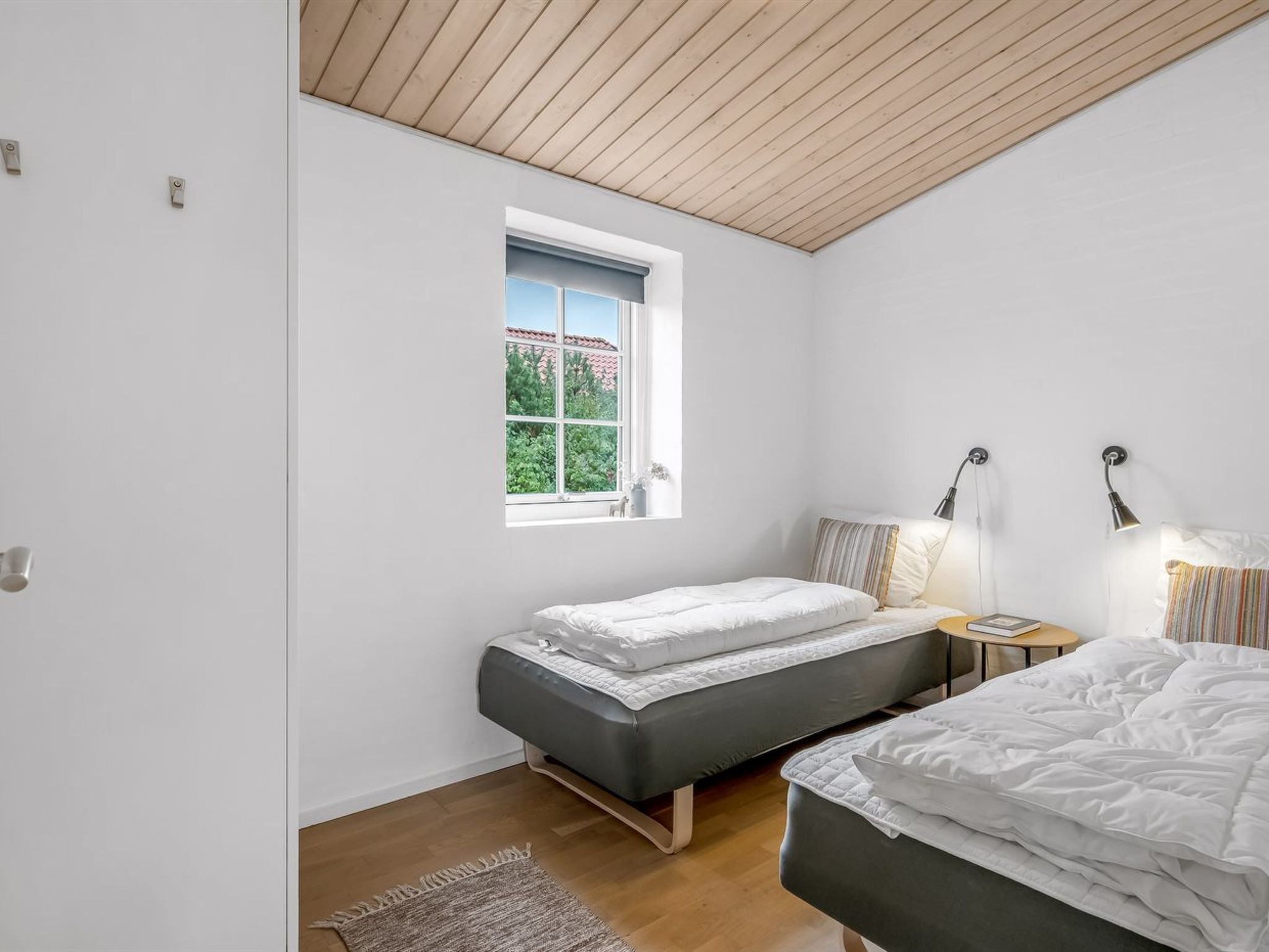 A bedroom with two beds, a window, a bedside table with a lamp, and a wooden ceiling.