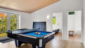 Ein moderner Indoor-Billardtisch mit blauer Spielbahnoberfläche, schwarzem Tischgestell und weißem Rand. Eine Tür führt in einen Raum mit einem Bett und einem kleinen Tisch. Der Raum hat weiße Wände, eine Holzbalkendecke und große Fenster mit Blick auf grüne Vegetation.