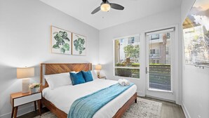Un dormitorio con cama de madera, ropa de cama blanca, manta azul, ventilador de techo y ventana con persianas.