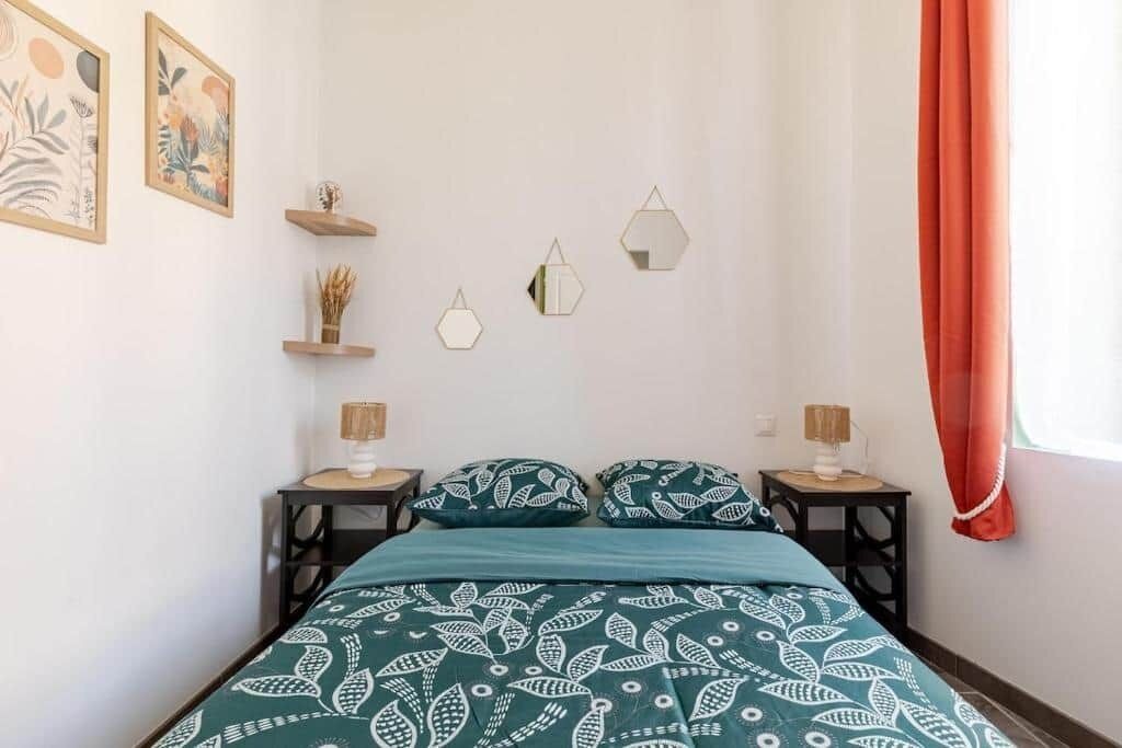 Ein Schlafzimmer mit einem Bett, zwei Nachttischen, einem Fenster mit Vorhängen und Wanddekorationen, darunter gerahmte Kunstwerke und geometrische Formen.
