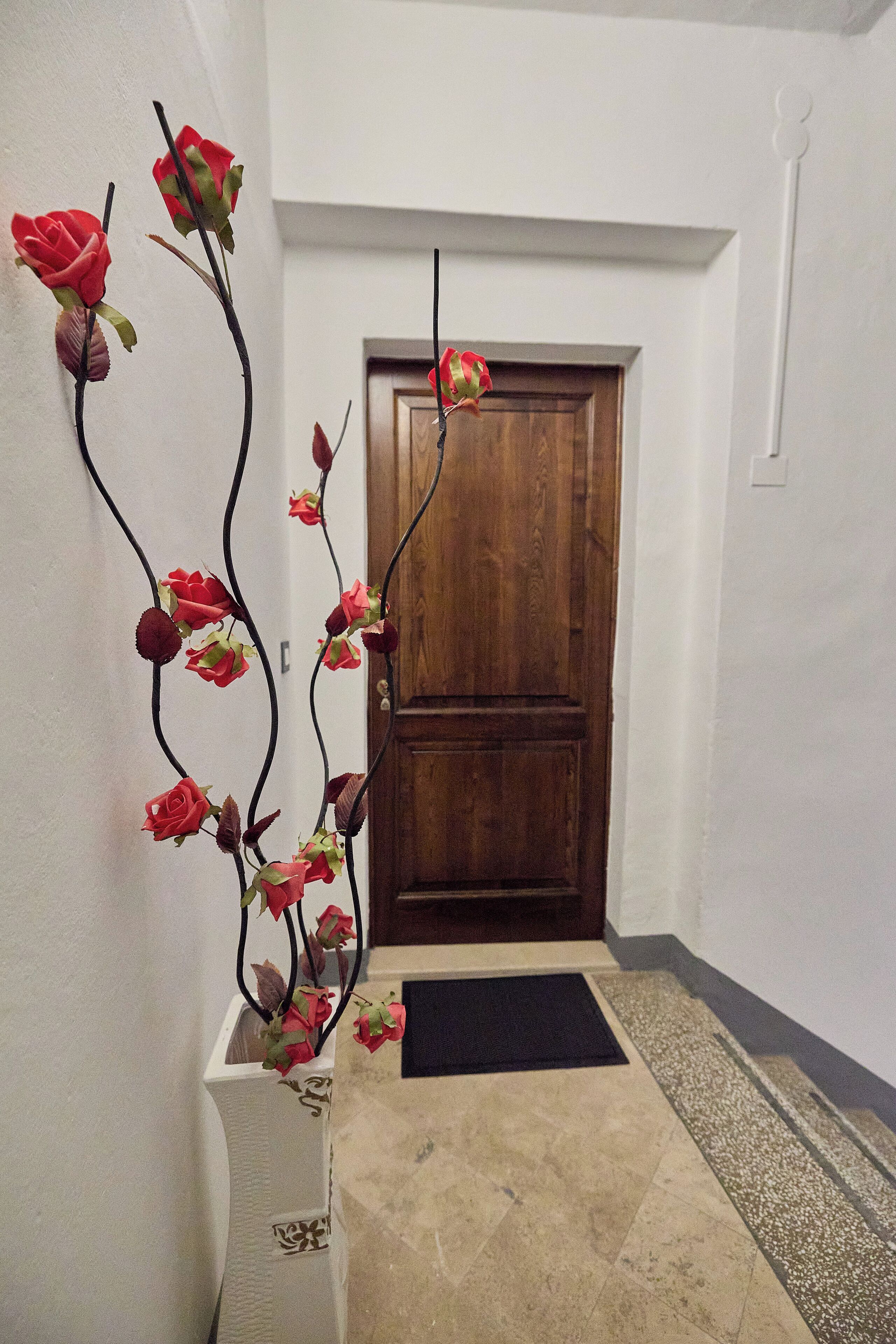 A hallway with a wooden door, a white wall, and a decorative metal plant with red flowers.