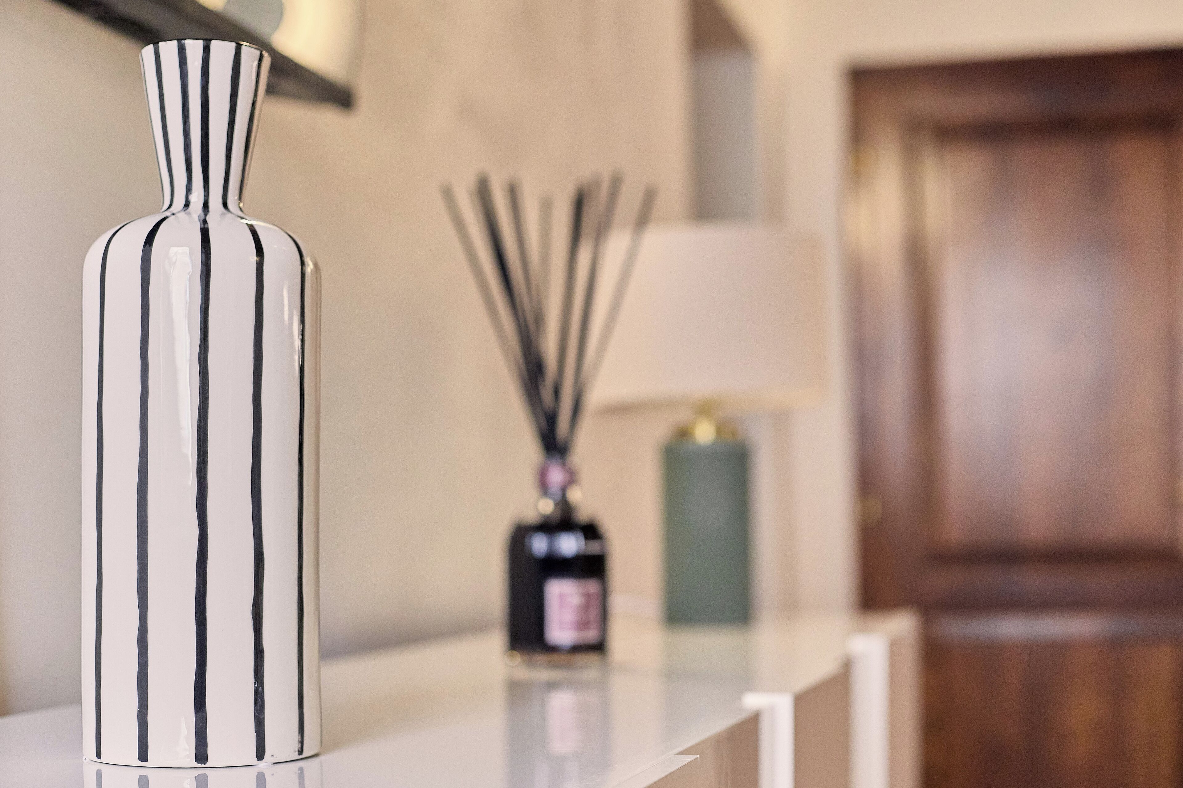 Un vase blanc à rayures noires, posé sur une surface réfléchissante, une bouteille noire avec une étiquette et, à l'arrière-plan, une lampe avec un abat-jour blanc.
