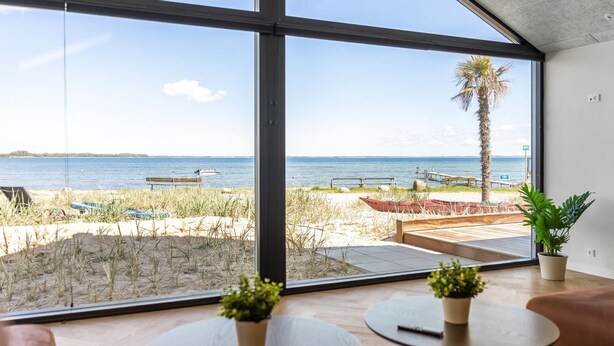 A modern room with large windows overlooking a beach, a wooden table, and a potted plant. - "Catalina" - 30m from the sea by Interhome (Sjølund)