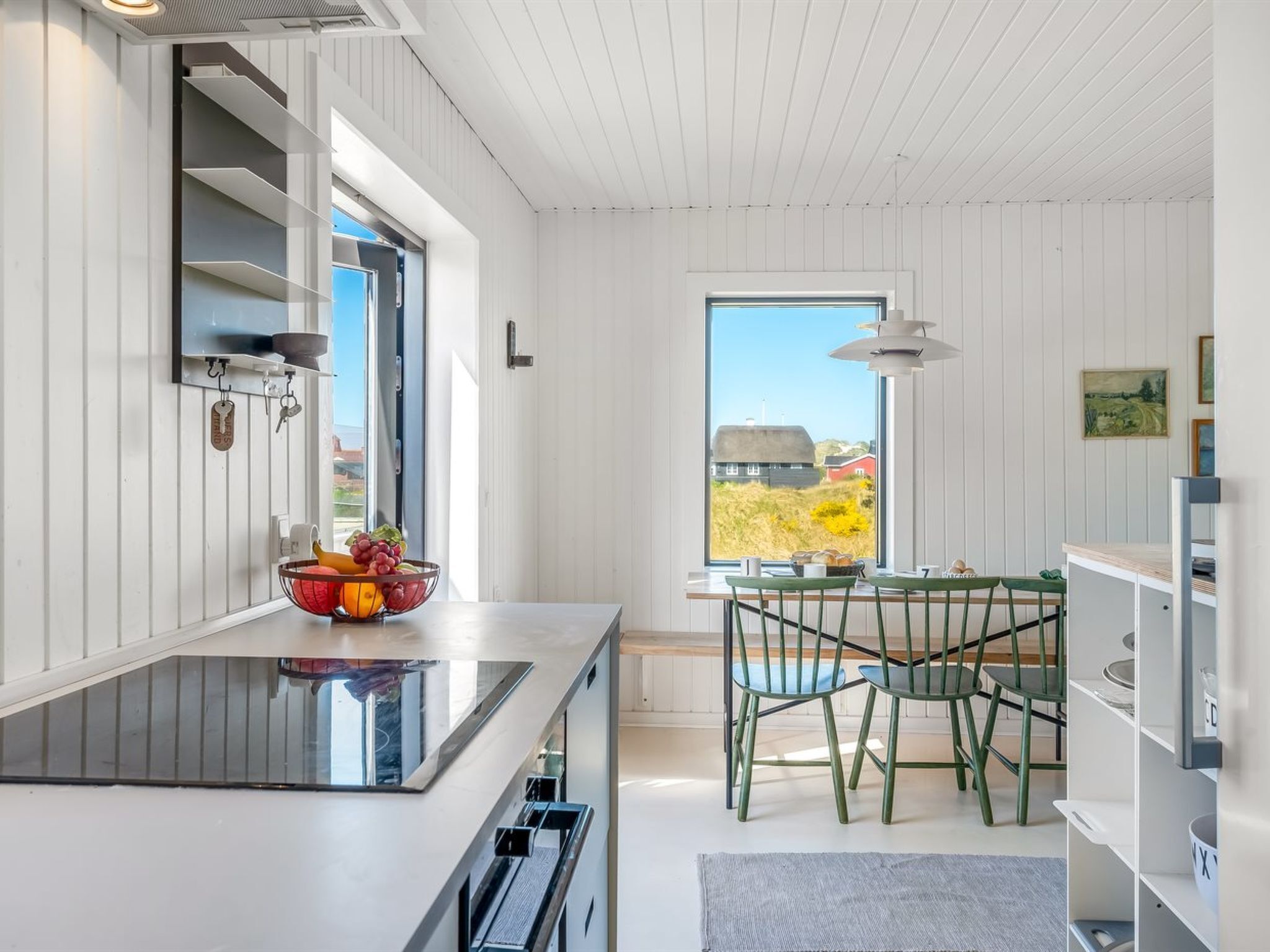 Eine moderne Küche mit großem Fenster, ein Essbereich mit grünen Stühlen und Blick auf einen roten Bauernhof und Felder.