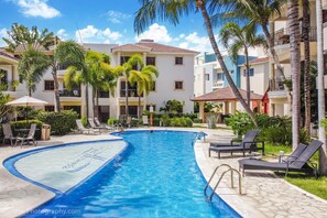 A swimming pool with lounge chairs and palm trees, surrounded by multi-story buildings with balconies. - Welcoming 2bedroom 2 bathroom apartment with AC ,pool, picnic area in Punta Cana (Punta Cana)