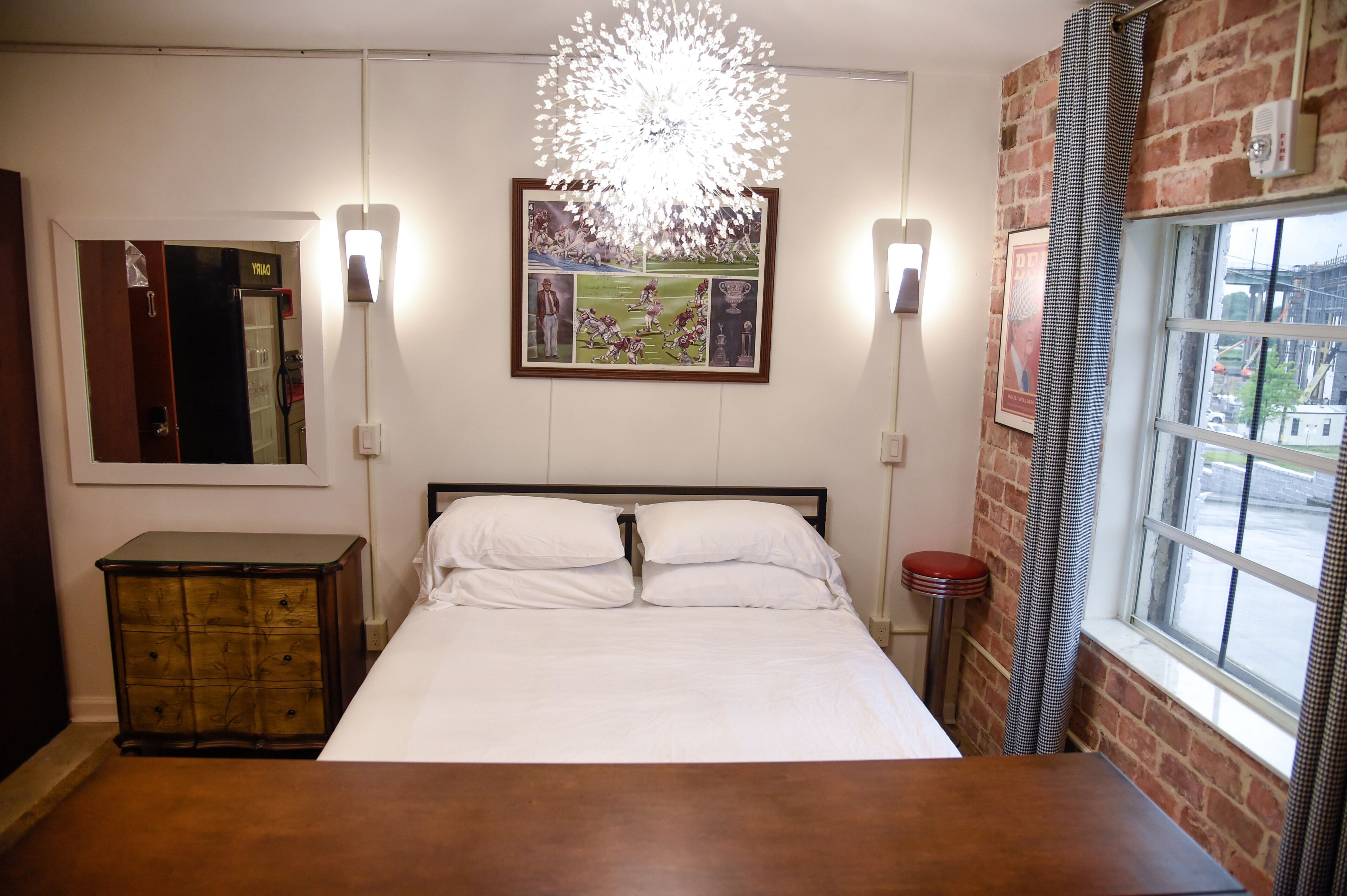A bedroom with a bed, a dresser, a wall-mounted light fixture, a framed picture, and a window with a view.
