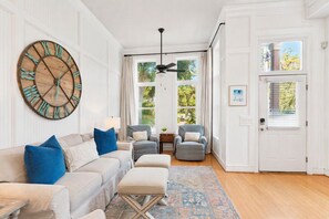 A living room with a large wall clock, a sofa with blue cushions, a grey armchair, and a patterned rug. - The Neptune House 3 bedrooms by the sea (St Simons Island)