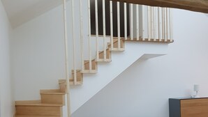 Uma escadaria com degraus de madeira e corrimãos brancos, um sofá cinzento e uma pequena mesa com uma luminária.