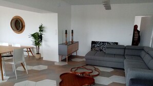Uma sala de estar moderna com um sofá seccional cinza, uma mesa de jantar de madeira com cadeiras, uma pequena mesa lateral e um espelho decorativo na parede.