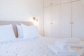 Ein ordentlich bezogenes Bett mit weißer Bettwäsche und einem Punkte-Muster, eine an der Wand befestigte Leuchte und eine Reihe weißer Schränke mit Messinggriffen.