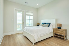 A bedroom with a white bed, wooden nightstand, and a view of the outdoors through a door. - Beautiful Second Row Beachside Home - Steps from Sand & Surf (Galveston)