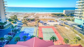 A view of a beachside tennis court complex with multiple courts, a parking area, and a building with balconies overlooking the beach.