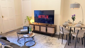 En stue med et fladskærms-tv der viser 'NETFLIX', et sofabord, en sofa, et spisebord med stole og en lampe.