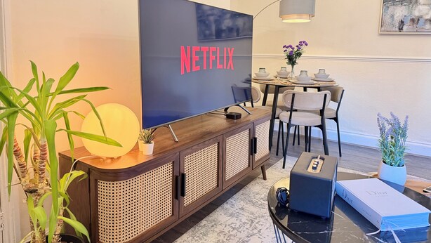 En moderne stue med et stort fladskærms-tv, der viser 'NETFLIX', et spiseområde med et rundt bord og stole, samt en dekorativ vase med blomster.