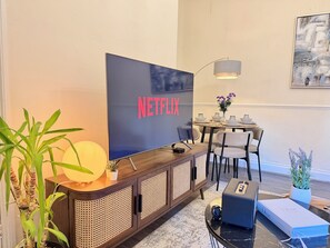 A modern living room with a large flat-screen TV displaying 'NETFLIX', a dining area with a round table and chairs, and a decorative vase with flowers. - 2BR Apartment, Fast Wi-Fi, Apple TV & Netflix, 20 Off Long Stays (Newcastle upon Tyne)