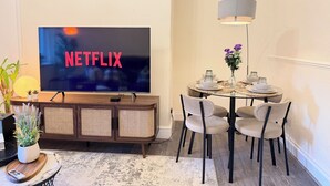 En moderne stue med et fladskærms-tv, der viser Netflix-logoet, et træskab, et spisebord med stole og en gulvlampe.