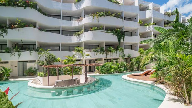 Ein mehrstöckiges Hotel mit Balkonen, die mit Pflanzen geschmückt sind, ein zentral gelegener Pool mit einer kreisförmigen Insel und Liegebereichen.