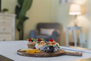Une planche en bois sur laquelle sont disposés des desserts et des biscuits, posée sur une table dans une pièce. On aperçoit une lampe et une chaise à l’arrière-plan.