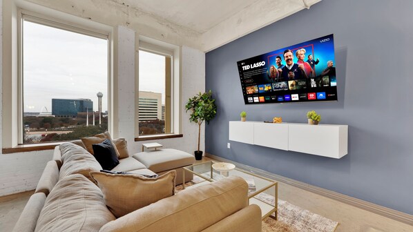 Una sala de estar moderna con un televisor grande montado en la pared, un sofá, una mesa de centro y vistas a un paisaje urbano a través de amplias ventanas.