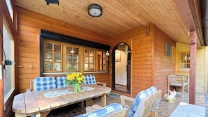 A wooden cabin with a dining area featuring a wooden table and blue plaid chairs, a large window with multiple panes, and a clock on the ceiling. - Vacation home in Wulsbüttel - Enjoy your vacation in Cuxland (Hagen im Bremischen)