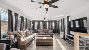 Amplia sala de estar con un sofá modular grande, reposapiés y un televisor de pantalla plana. Se observan ventiladores de techo, ventanas grandes con cortinas y una pared de ladrillo al fondo.