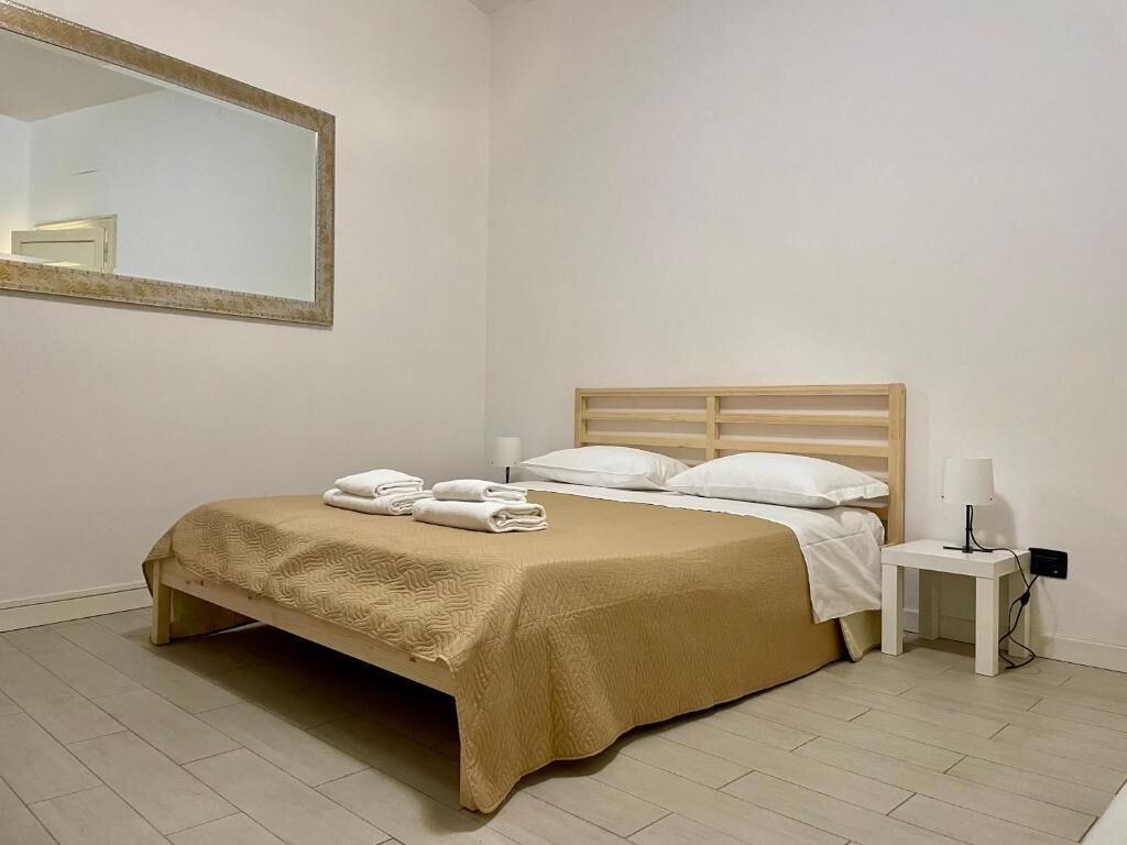 A neatly made bed with a beige bedspread and white pillows, a bedside table with a lamp, and a large mirror on the wall.
