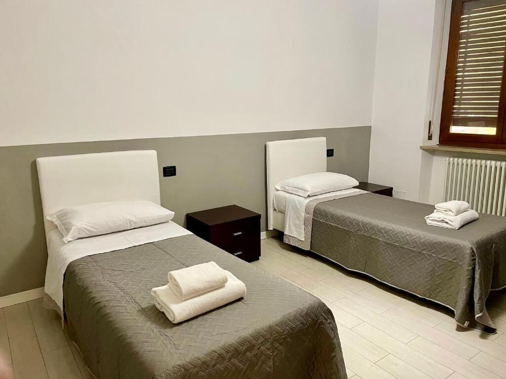 A hotel room with two single beds, each with a pillow and a folded towel. There is a small bedside table between the beds.