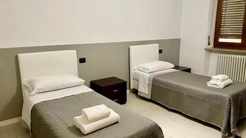 A hotel room with two single beds, each with a pillow and a folded towel. There is a small bedside table between the beds.