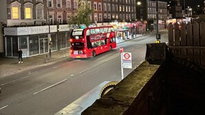 Scena notturna di una strada con un autobus a due piani rosso, edifici con vetrine e un cartello stradale.
